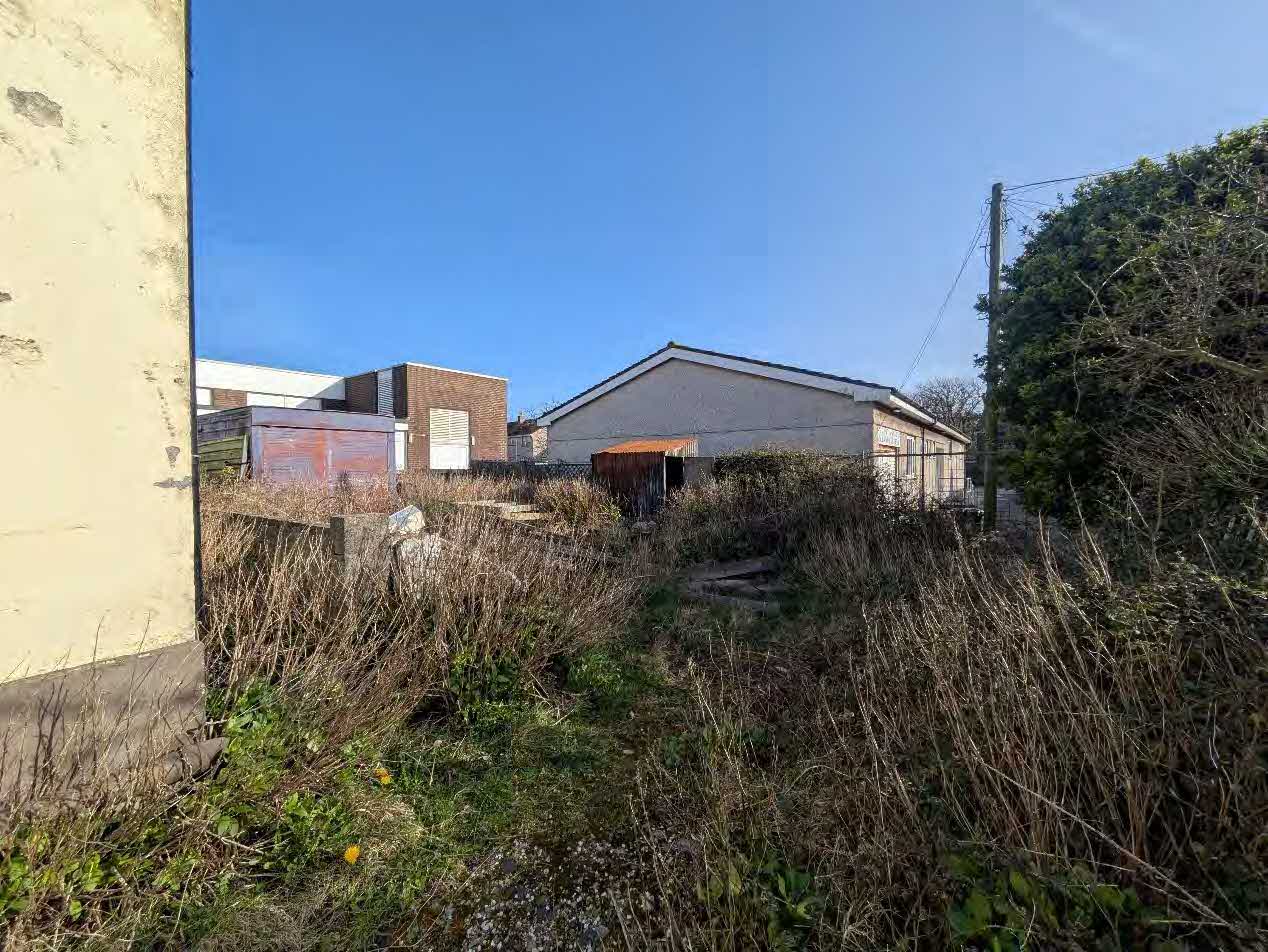 A photograph showing an overgrown plot of land with existing outbuildings and structures in the background under a clear blue sky.
