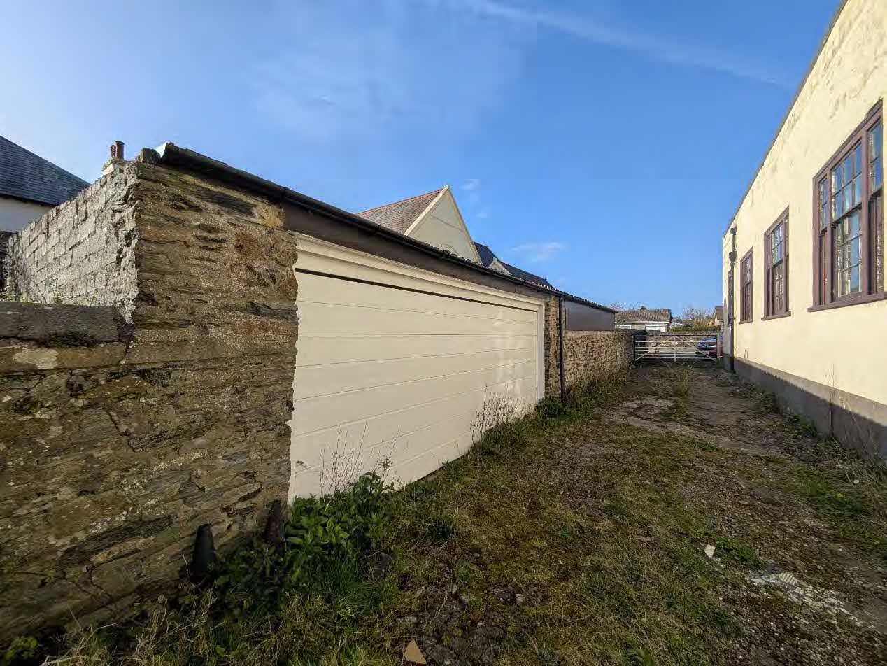 A photograph showing a narrow grassy path between a white garage structure with an old stone wall and a cream-colored house.