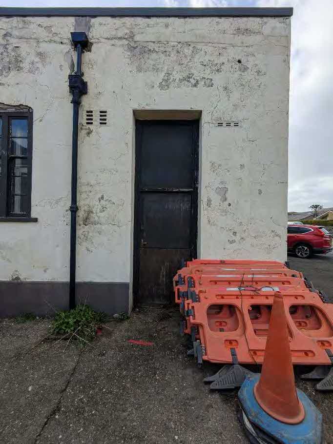 A weathered white building exterior featuring a dark door and window, with stacked orange traffic barriers and a cone in the foreground.