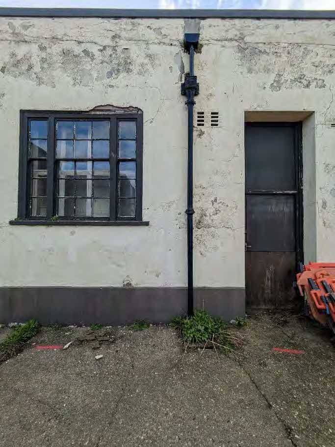 A photograph showing the weathered exterior wall of a single-story building featuring a black-framed window, a vertical downpipe, and a dark door.