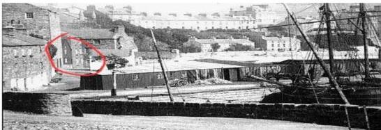 A black and white photograph showing a coastal harbor scene with a specific stone building circled in red in the middle ground.