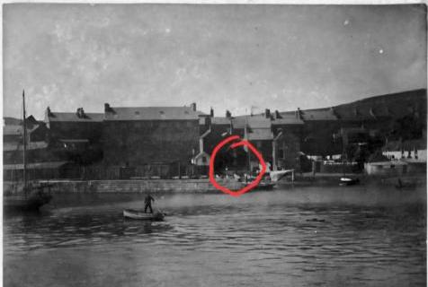 A black and white historical photograph showing a row of terraced buildings along a waterfront with a small boat in the foreground.