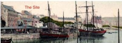 A photograph showing a harbor scene with several sailing ships docked near a waterfront lined with buildings. The text 'The Site' is overlaid in red, indicating the location context.