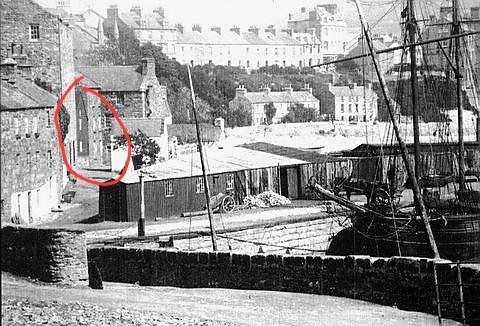 A black and white photograph showing a coastal harbor scene with a red circle highlighting a stone building, set against a backdrop of terraced houses on a hillside.