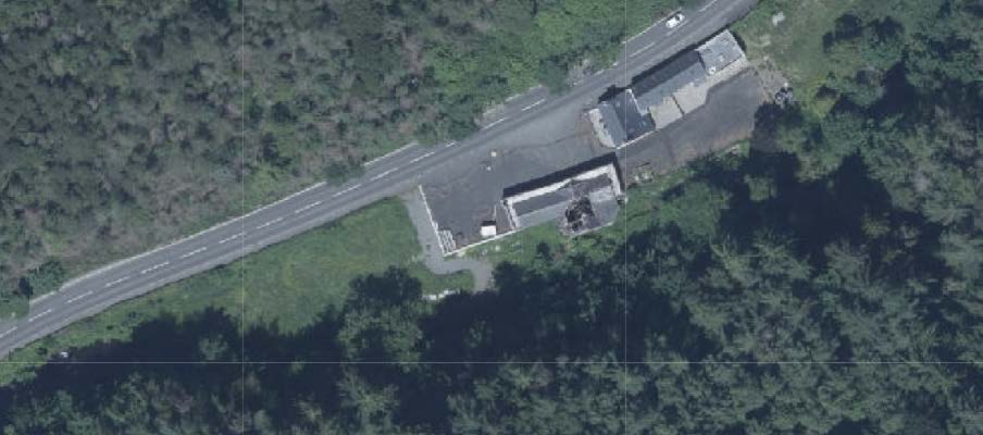 Aerial satellite view of a site adjacent to a road showing buildings, paved areas, and surrounding vegetation.