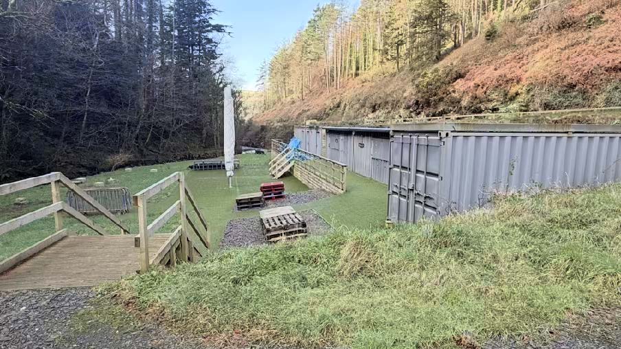 A photograph showing a row of grey shipping containers situated on a grassy slope with wooden decking, stairs, and artificial grass.