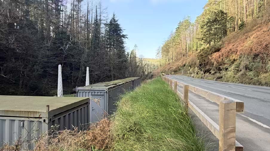 A roadside view showing several grey shipping containers situated next to a wooded area, separated from the road by a grassy verge and wooden fence.