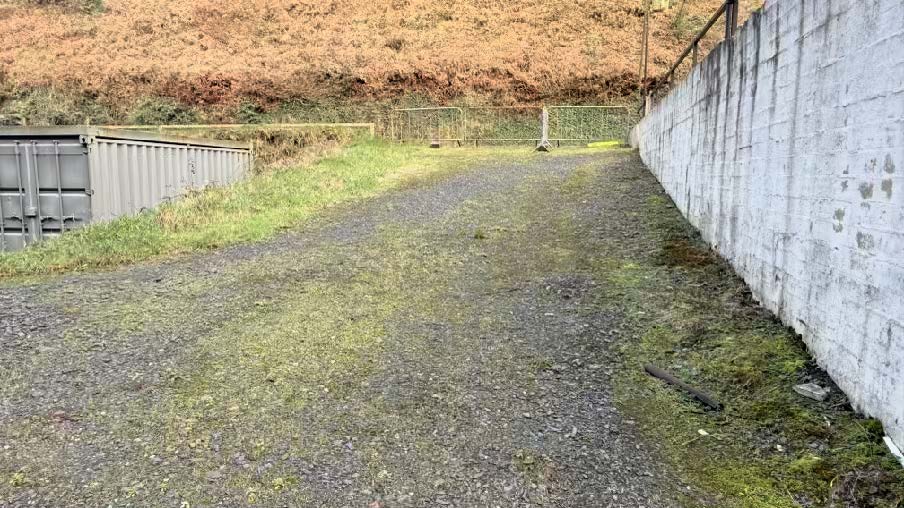 A photograph showing a gravel access track running alongside a tall white concrete wall on the right and grey shipping containers on the left.