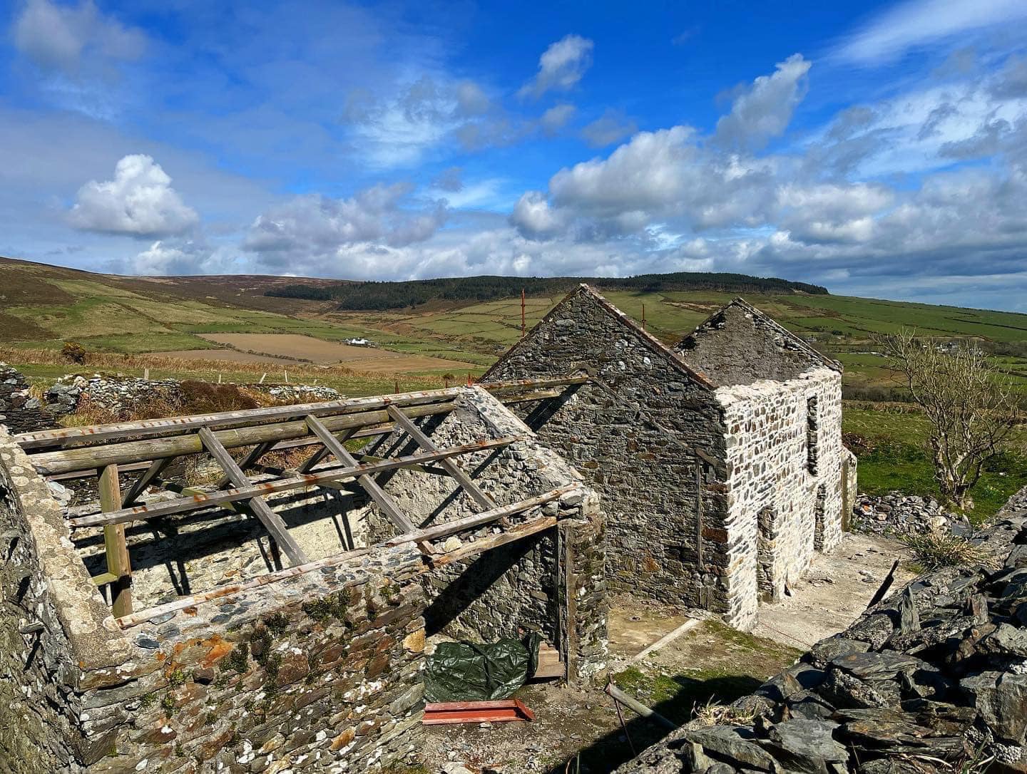 A photograph showing a cluster of old stone buildings in a rural setting, including a roofless structure with exposed timber framing next to two other stone buildings.
