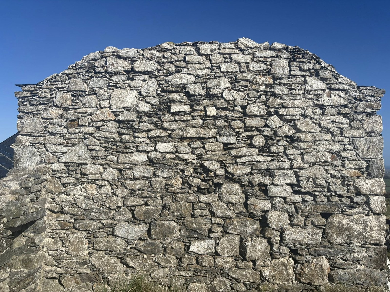 A close-up photograph of a rough stone gable wall, likely part of an existing agricultural building, set against a clear blue sky.
